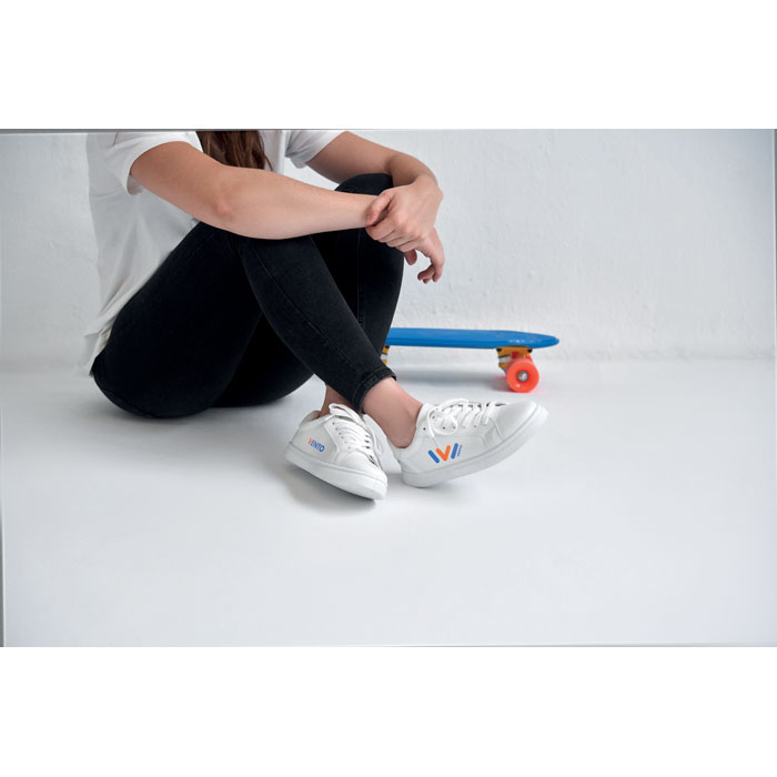 Vista parcial de una persona sentada con vaqueros negros, zapatillas blancas y un monopatín azul apoyado en el suelo.